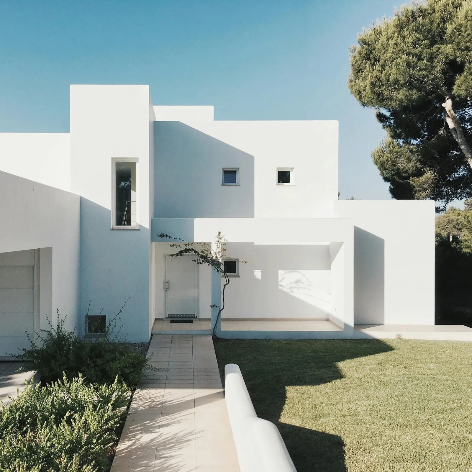White cubic house with a garden path and tall pine trees