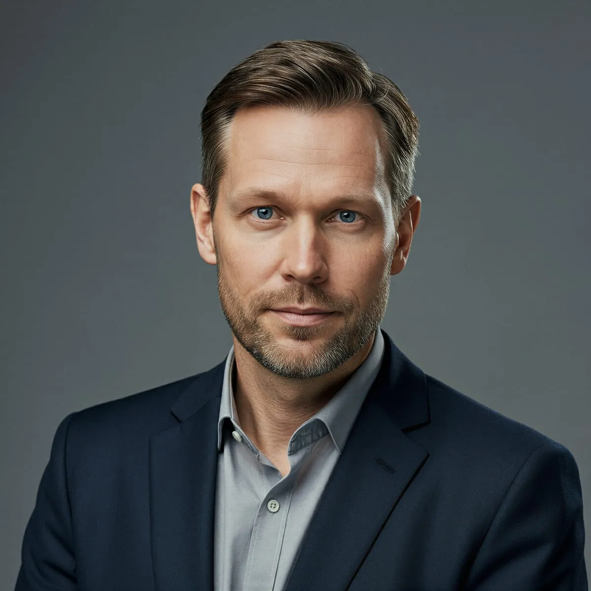Man with short brown hair and a light beard in a navy blazer and grey shirt